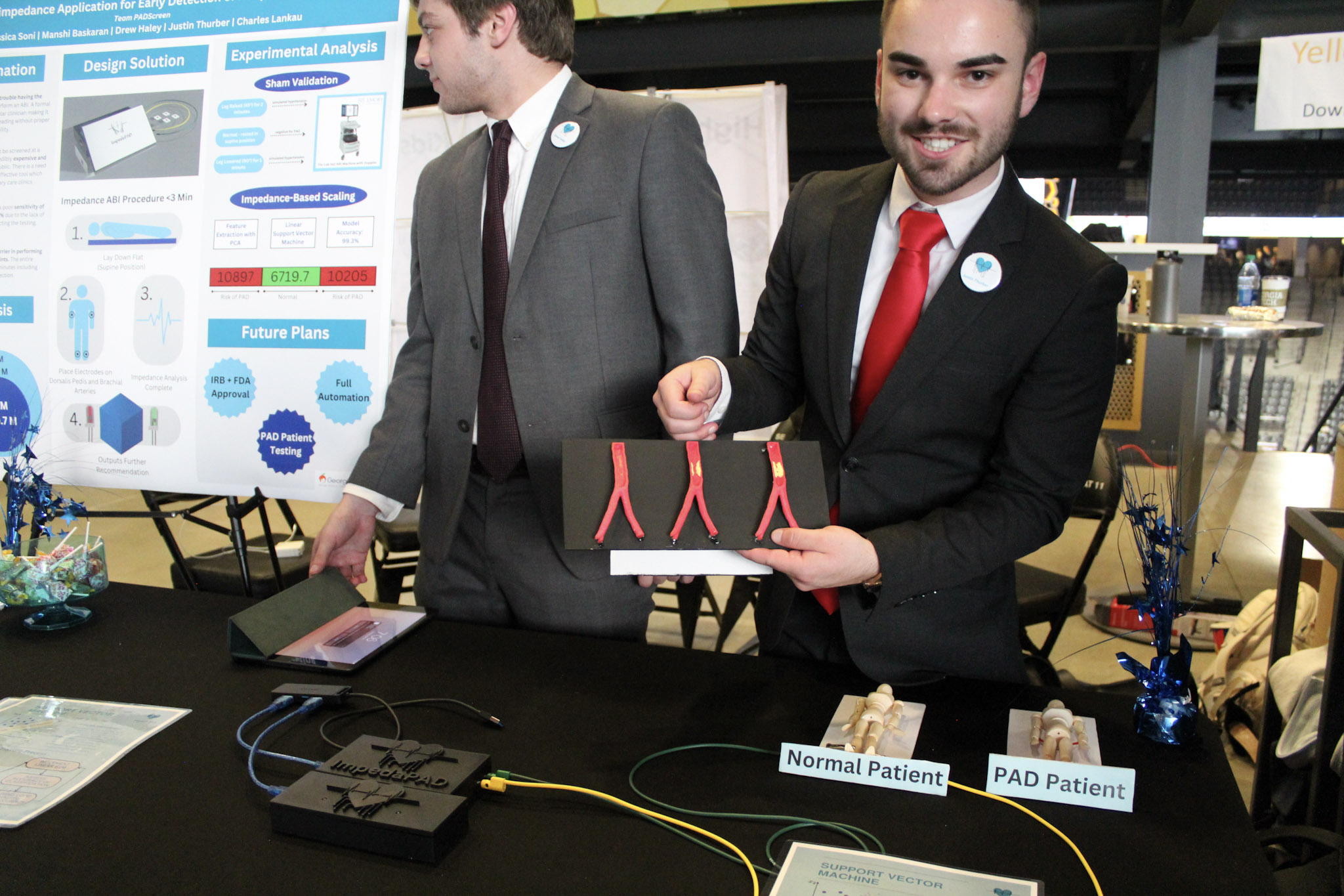 A male student shows a model of three different stages of arterial disease as part of his team's capstone project. Another male student stands behind him near a poster presentation board.