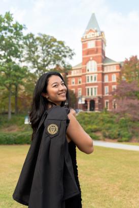 Photo of Sierra Venetta standing on a grassy area of the Georgia Tech campus with the Tech Tower building in the background. She has her back facing the viewer and her graduation gown is draped over her shoulder and she looks back toward the viewer.