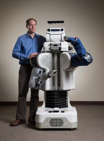 The blue cover on the arm of this robot protects an array of sensors that help it reach through the clutter of a typical home to perform personal care tasks. Charlie Kemp poses with the robot, which also holds an electric shaver. (Credit: Rob Felt)