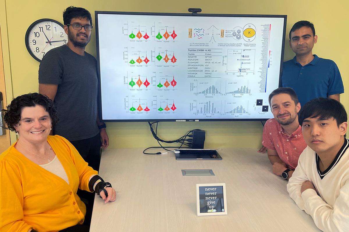 Cassie Mitchell at a table with her Ph.D. students. On the screen above the table is a data slide showing their work on an integrative machine learning algorithm to predict risk of neurodegenerative disease progression based on patient proteins, genotype, demographics, cognitive testing, and medical history. Clockwise from top left are Irfan Al-Hussaini , Raghav Tandon, David Kartchner, Albert Lee, and Mitchell. (Photo Courtesy: Cassie Mitchell)