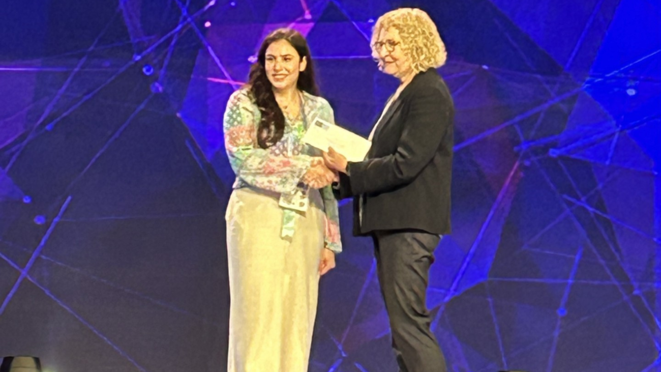 Close up photo of Retta El Sayed dressed in white standing on a stage being given an award by a female presenter.