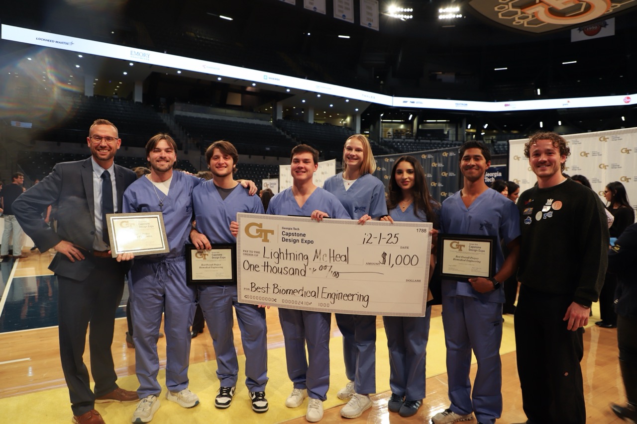 A photo of a team of students in blue scrubs standing in front of a step and repeat banner holding a big check award while their faculty advisors stand to either side of the group.