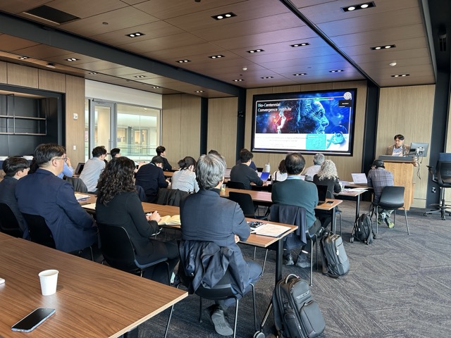 Photo shot from the back of a room where a group of people sitting at long tables look toward a projection screen for a presentation. Presenter in the front right side standing at the podium.