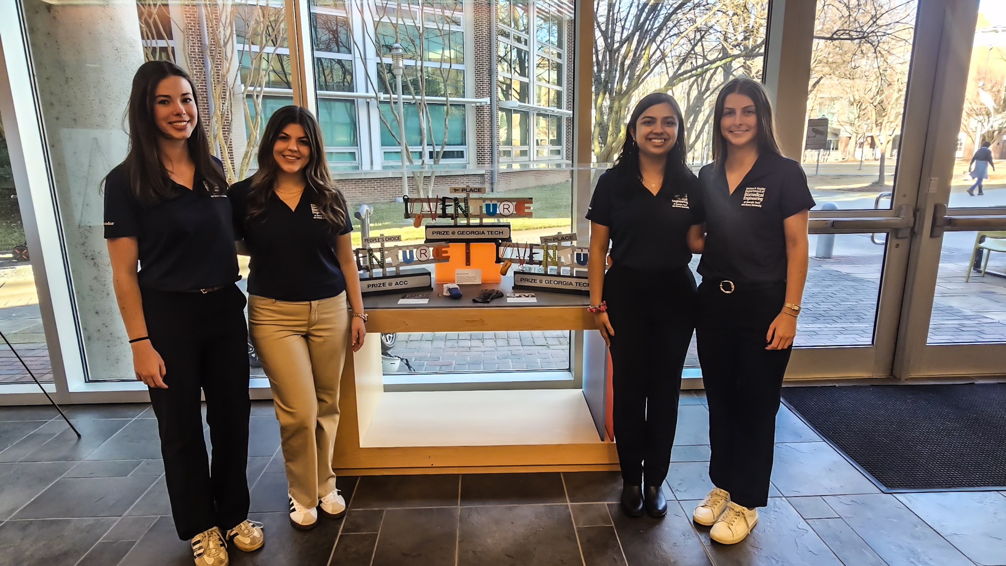 Group photo of four female students who are on the BMED Ambassadors team. Names are unkown. The women flank a display case with the Georgia Tech Inventure Prize trophy.