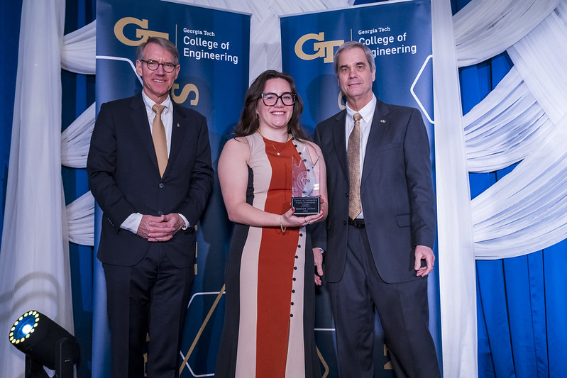 Three people pose on a stage with Georgia Tech College of Engineering banners; the person in the center holds a glass award.