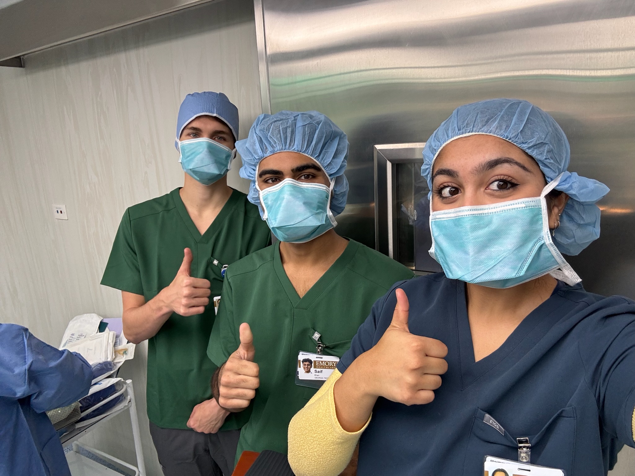 Three individuals in surgical scrubs and hair covers stand together in a clinical setting, each giving a thumbs‑up gesture. Medical equipment and stainless‑steel surfaces are visible in the background.