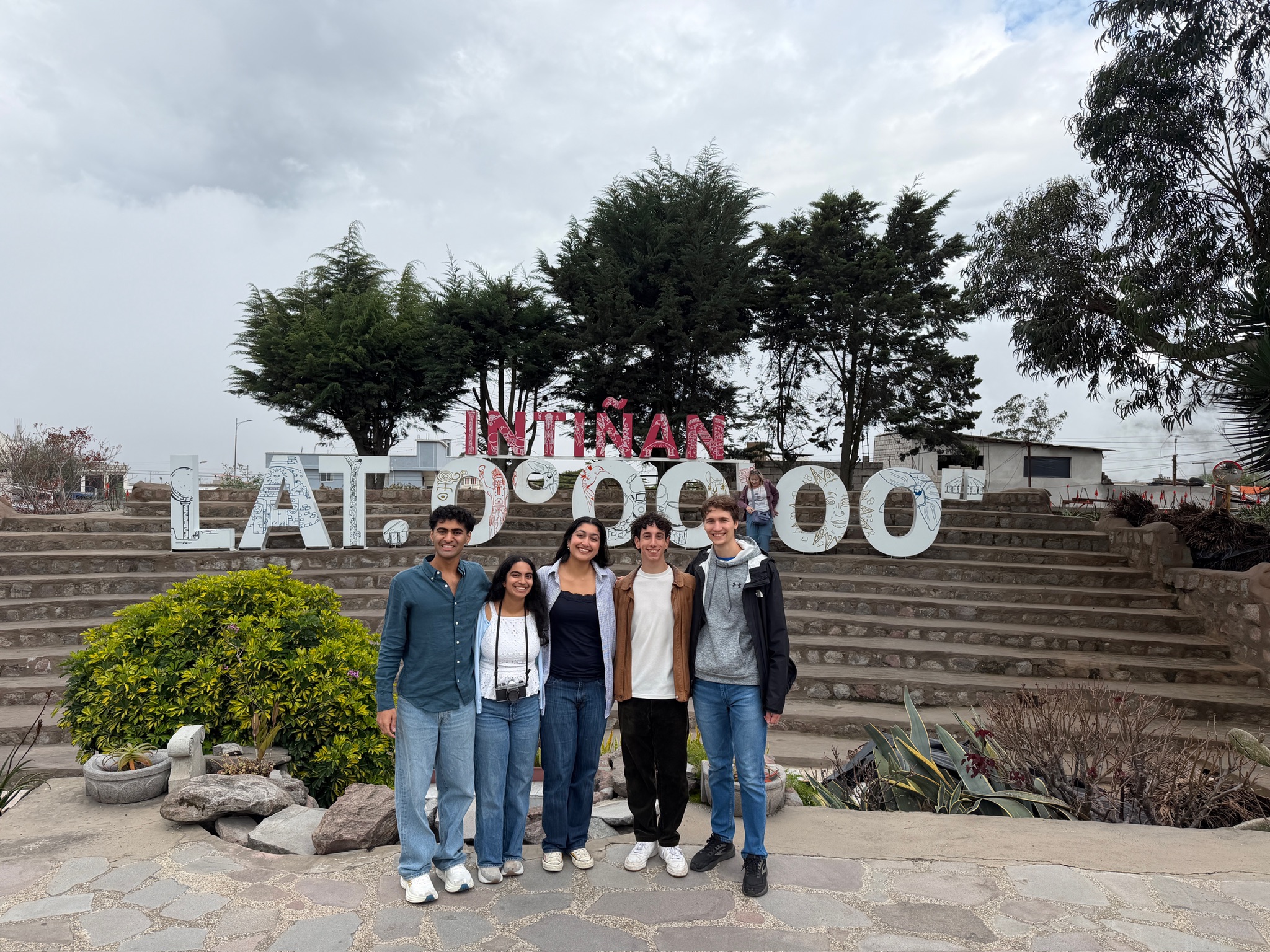 A group of five people stands together in front of a large outdoor display at the Intiñan Museum in Ecuador, featuring big white letters marking latitude 0°0'0". The group is positioned on a stone walkway with steps and tall trees in the background, under a cloudy sky.