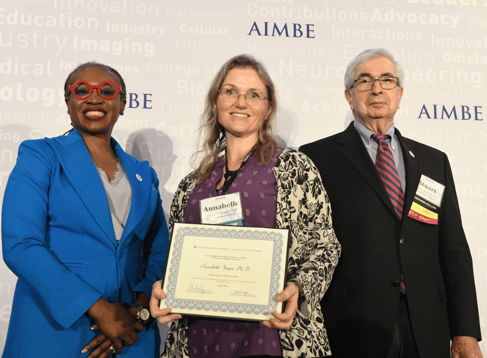 Three individuals standing in front of an AIMBE backdrop; the center person holds an AIMBE College of Fellows certificate during an award photo.