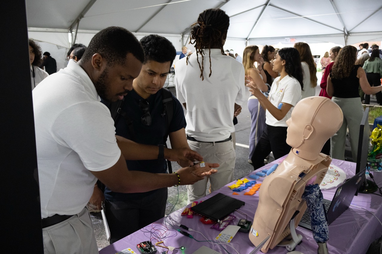 An African American male student on a BME capstone team shows a Capstone Expo attendee (male) the device his team created. Other students are wlaking around in the background inside of the large tent exhibit space.