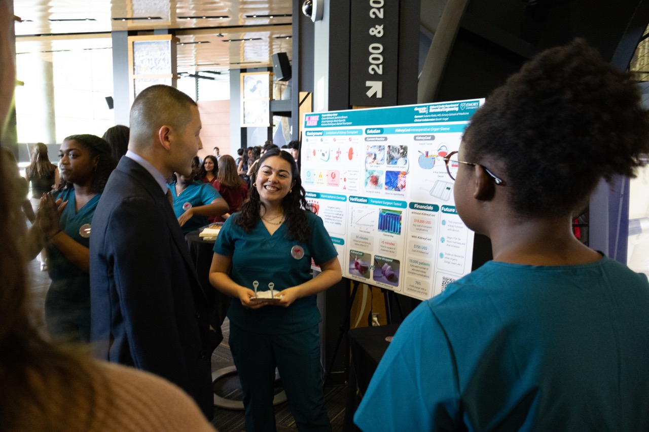 Two women members of Team Kidney Krew show a Capstone Expo attendee (male) their project.