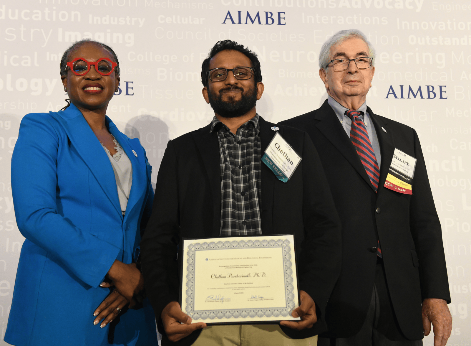 Three individuals standing in front of an AIMBE backdrop; the center person holds an AIMBE College of Fellows certificate during an award photo.