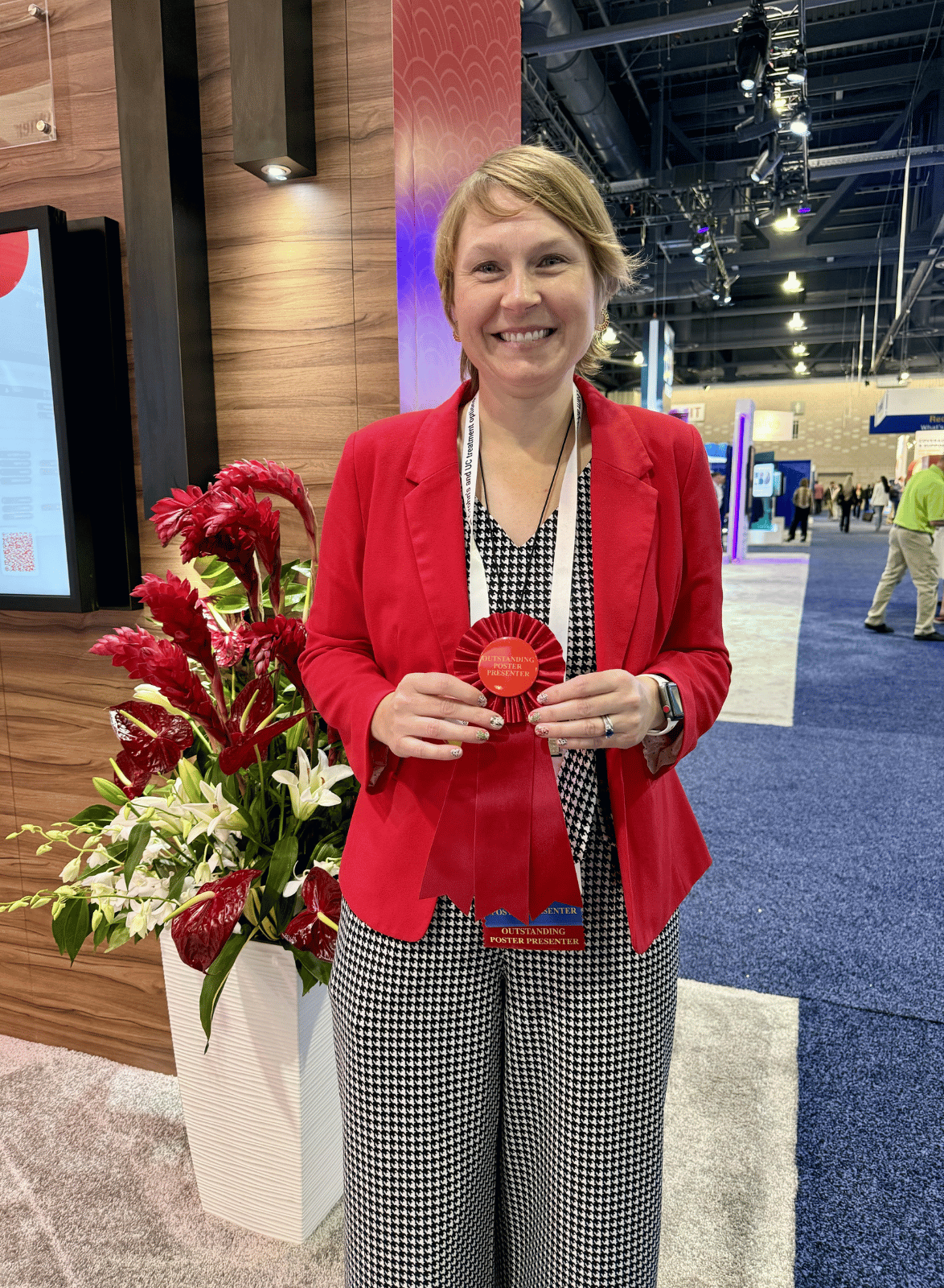 Person standing in a conference hall holding a red rosette ribbon labeled “Outstanding Poster Presenter,” with a scientific meeting floor visible in the background.