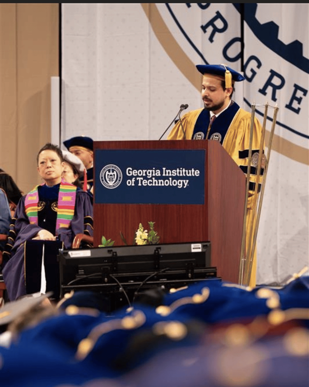 Individual in academic regalia speaks at a podium labeled “Georgia Institute of Technology,” with faculty seated behind and a blurred audience in the foreground.