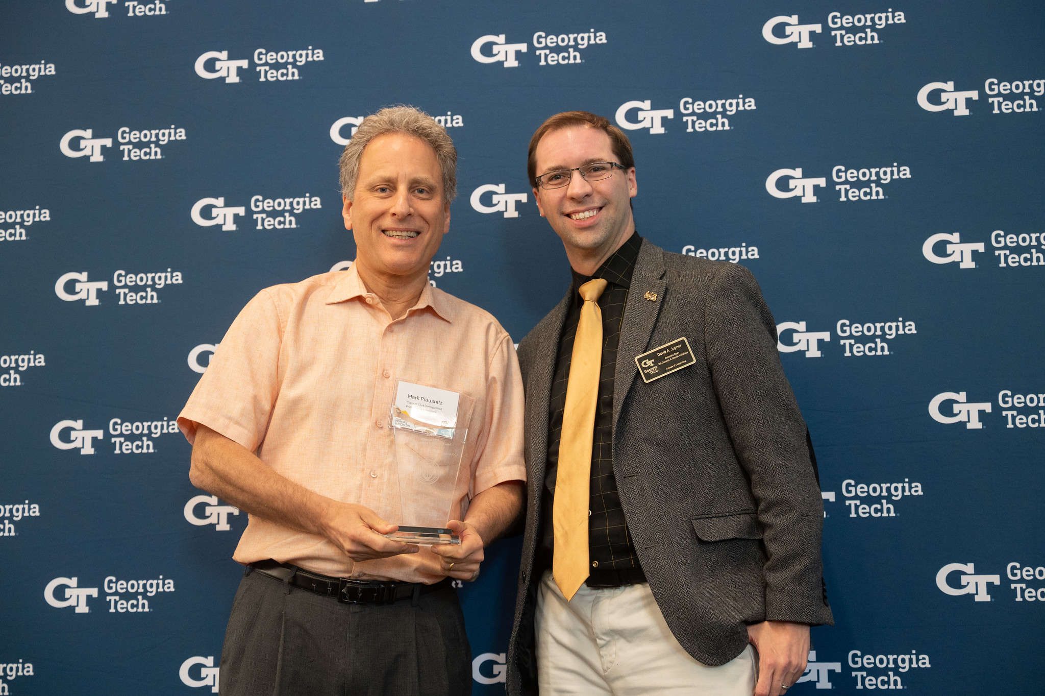 Two individuals pose in front of a Georgia Tech backdrop, with one holding a clear glass award.