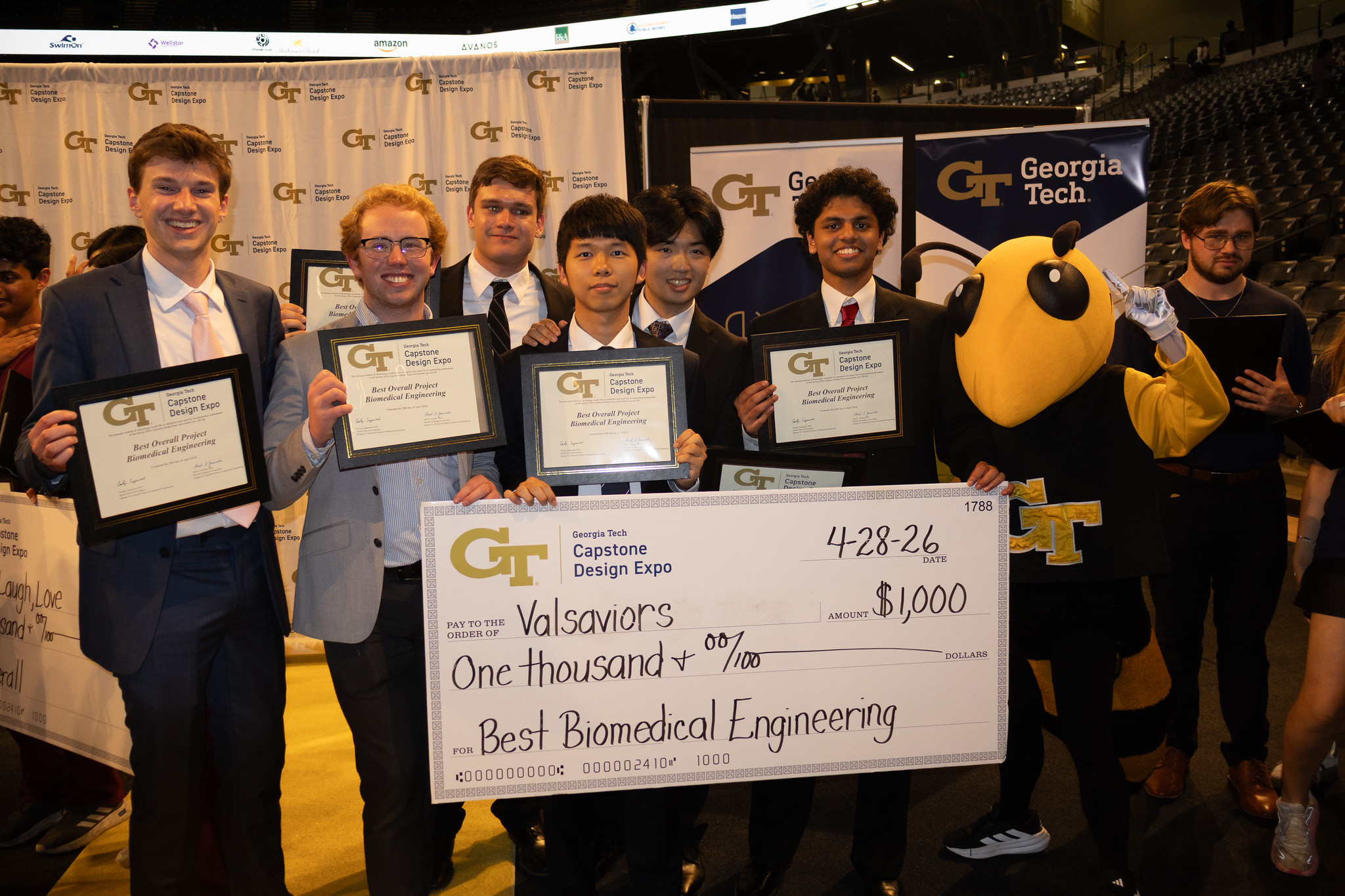 Group of students holding certificates and a large $1,000 check for “Best Biomedical Engineering” at the Georgia Tech Capstone Design Expo, standing in front of Georgia Tech banners, with the Buzz mascot beside them.