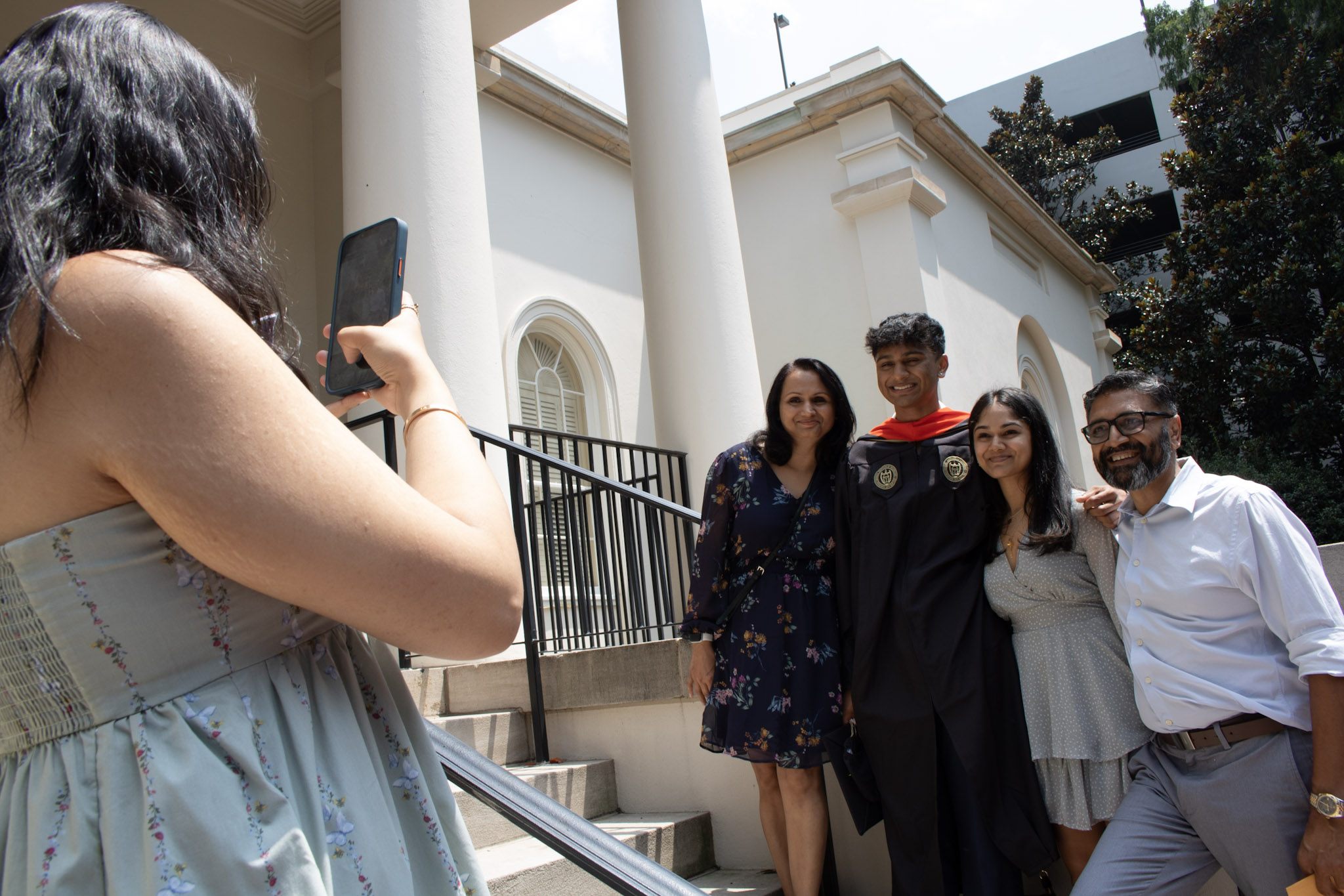 A young woman takes a photo with her cell phone of a male graduate of the MBID program standing with his parents.