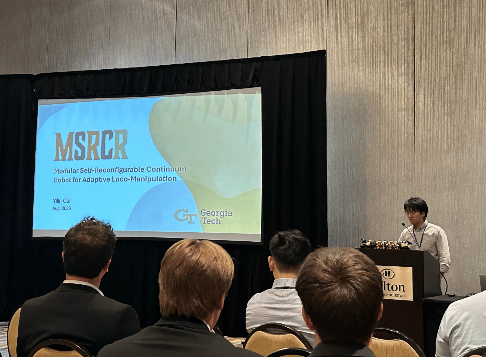 A researcher presents a talk at a conference, standing at a podium beside a projected slide titled “MSRCR: Modular Self-Reconfigurable Continuum Robot for Adaptive Loco-Manipulation.” Audience members are seated facing the screen, and a small prototype robot rests on the podium during the presentation.