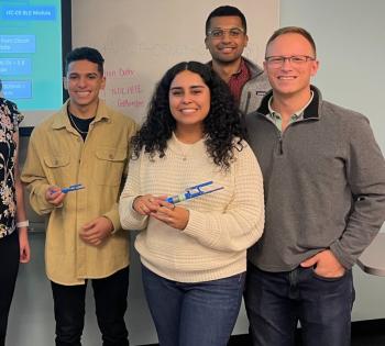 A photo of a group of individuals. From left to right: a male student, a female student, and a male professor. Another male student stands behind the female student.