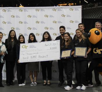 Photo of a group of nine students representing teams AdvanCED and Jawws holding a check for $1000. Two female students on the right side of the photo are holding small plaques. Next to the students on the far right of the photo is the Buzz mascot and Dr. James Stubbs, faculty advisor for BME Capstone.