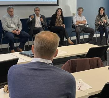 Photo of five McCamish conference panelists. Four panelists (one man and three women) listen to the fourth participant, a male, speaking.