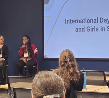 Photo of three women sitting at the front of a meeting room with a large screen that says "International Day of Women and Girls in Science." One woman who is African American, holds a mic while she is speaking.