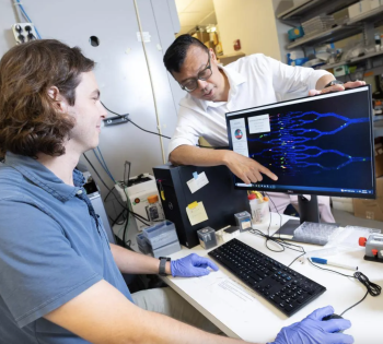 A male student with a blue shirt sits in front of a computer moving the computer mouse as a male faculty member, standing behind the computer screen, points to a digital DNA sequence on the screen.
