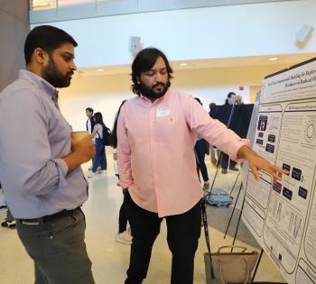 Photo of male student in a pink shirt in the center of the photo pointing to a poster of his research as another male student in a blue shirt on the left side of the photo looks on.