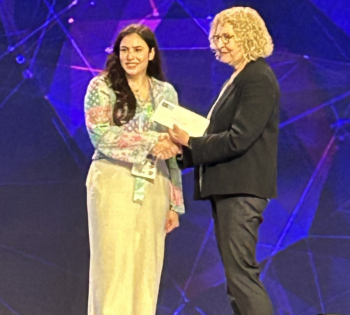 Close up photo of Retta El Sayed dressed in white standing on a stage being given an award by a female presenter.