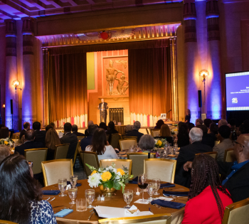 Photo of the Egyptian Ballroom at the Fox Theatre Atlanta looking toward the stage. In the foreground are people sitting in banquet chairs at tables looking at the speaker. In the background is the stage and speaker.