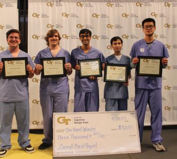 Five male students from Capstone team One-Hand Wonder holding gold plaques for winning first place at Capstone Expo. A large check fro $3,000 sits on the floor in front of them.