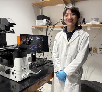Photo of Zhou Fang in lab standing next to laboratory equipment wearing PPE.
