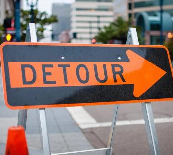 Photo of a detour road sign that has an orange border and an orange arrow with the words "DETOUR" in black inside of the arrow