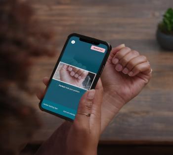 Photo of an over the shoulder shot of a cell phone being held by a woman. She uses the phone to take a photo of her fingernails.