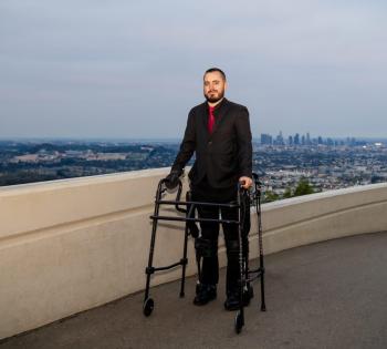 Photo of Coulter BME Ph.D. student Ignacio Montoya, standing on some sort of building rooftop with the skyline of Los Angeles behind him in the background. He is standing with the use of a walker.