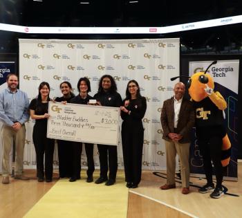 A photo of a team of students in black scrubs standing in front of a step and repeat banner holding a big check award while their faculty advisor stands to the left of them and the Georgia Tech Buzz mascot stands to the right of them next to the provost.