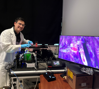 A person wearing a white lab coat and blue gloves is working at a laboratory station equipped with advanced imaging equipment. The setup includes multiple tubes, wires, and containers connected to a black optical device. A large monitor on the right displays a vivid, high-resolution microscopic image in shades of purple and pink, showing tissue structures. 