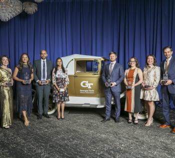 A group of people stand on a stage in front of a vintage Georgia Tech Ramblin’ Wreck car, each holding a glass award, with blue draping and chandeliers in the background.