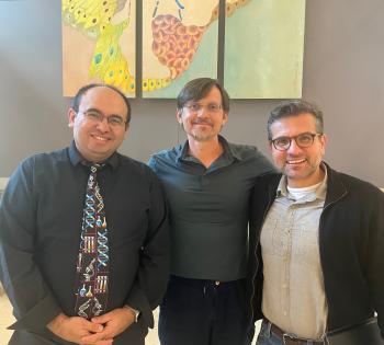 Three people stand side by side indoors for a group photo, with colorful scientific artwork displayed on the wall behind them
