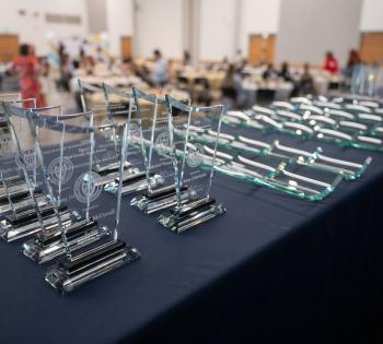 A row of clear glass award plaques arranged on a table in the foreground, with more plaques laid flat beside them and a blurred event setup in the background.