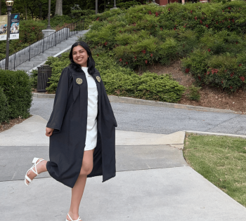 A graduate in cap and gown stands on the Georgia Tech campus with Tech Tower rising in the background, surrounded by trees and landscaped walkways.