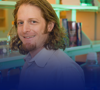 Smiling person with shoulder-length hair posing in a laboratory setting, with shelves of glass containers and equipment visible in the background.