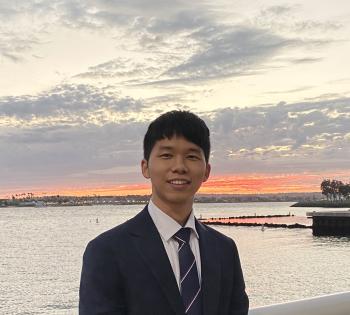 Person in a suit standing by the water at sunset, with a cloudy sky and shoreline in the background.