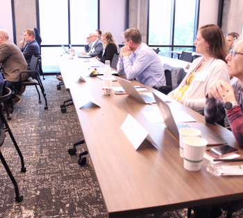 Audience members sit around a conference table in a bright meeting room, listening to a speaker holding a microphone; laptops, notebooks, and coffee cups are visible on the table near large windows.
