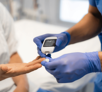 Healthcare worker wearing blue gloves uses a handheld glucose meter to check a patient’s blood sugar from a finger prick.