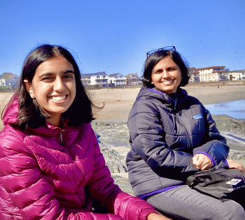 Two people in puffer jackets sit on rocks at a beach with beachfront homes and a clear blue sky in the background.