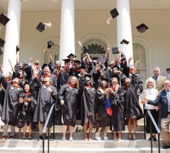 Graduates of the MBID program in Coulter BME are standing on the steps of the Historic Academy of Medicine with their caps and gowns on tossing their caps in the air. The department chair, two faculty, and the program director or standing on the right side of the steps. 