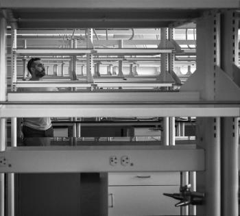 A person stands between rows of laboratory benches and shelving in a black-and-white research lab photograph.
