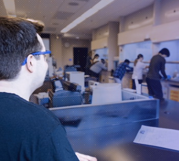 A student wearing safety glasses works in the foreground of a robotics lab as several other students work at equipment in the background.