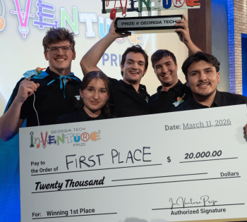 Five members of the winning Georgia Tech InVenture Prize team pose onstage holding a large ceremonial check for $20,000 and a trophy labeled “Prize @ Georgia Tech.” The oversized check reads “First Place” and “Winning 1st Place,” with the Georgia Tech InVenture Prize logo displayed prominently in the foreground. Colorful InVenture Prize signage appears in the background.