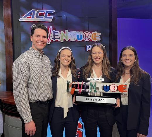 Photo of three female students and their mentor, a male, taking a group photo holding one of the Inventure Prize award trophies for winning at the competition for their medical device